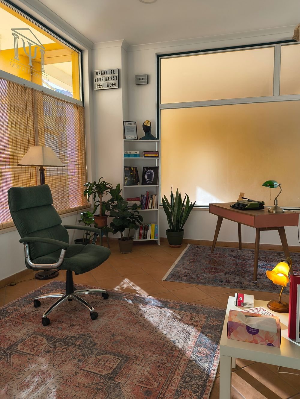 Sala de consulta com cadeirão verde, plantas e estante, com luz natural